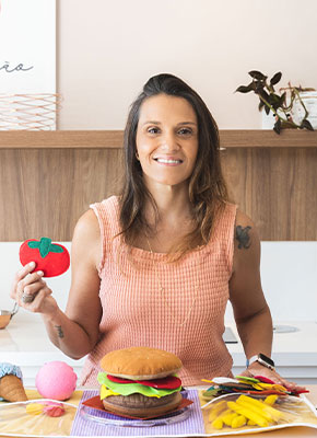 Renata Riciati segurando um tomate de brinquedo, com um hambúrguer de feltro sobre a mesa